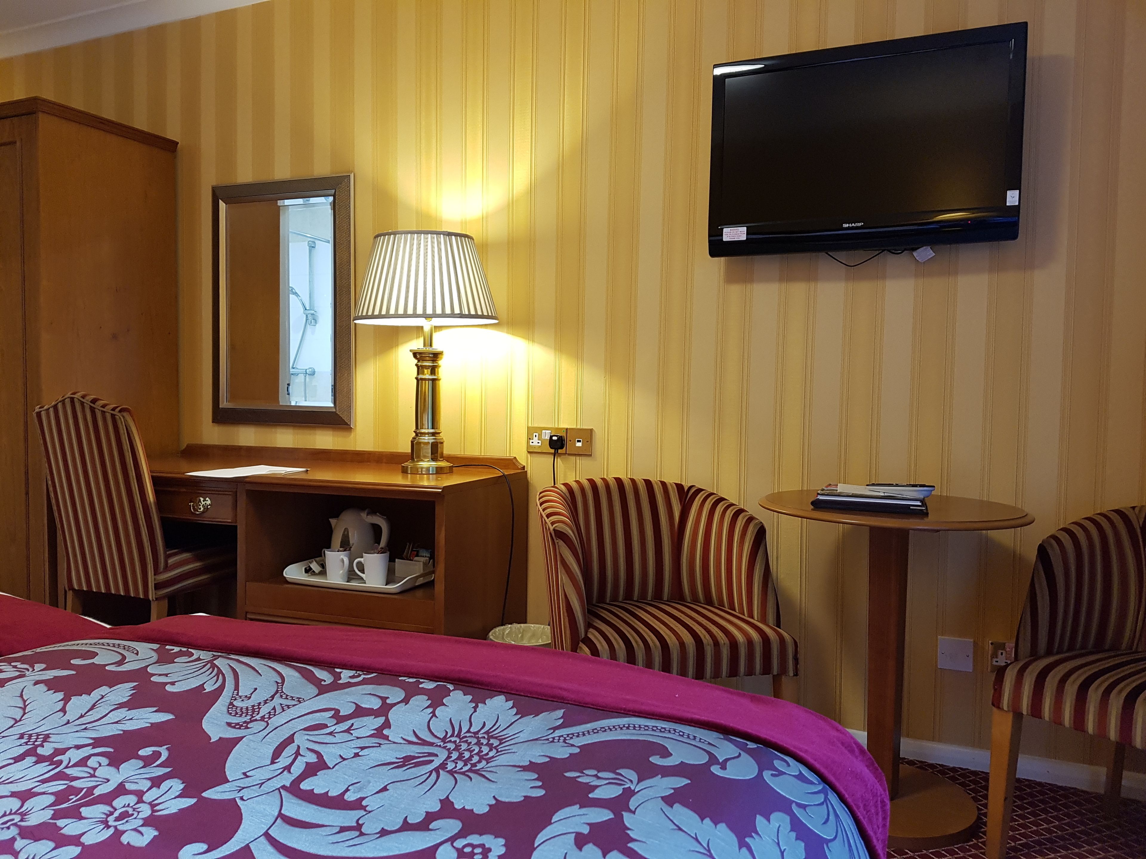 A hotel room with a bed, a desk with a lamp, two striped chairs, a small table with a book, a TV mounted on the wall, and a mirror.