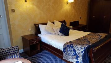 A hotel room with a large bed, two bedside tables, a nightstand, a chair, and a small table with a remote control and books.