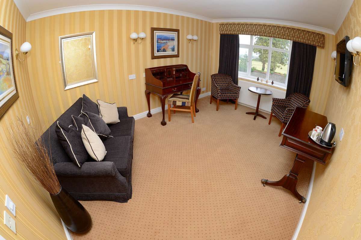 A room with a dark-colored sofa, a wooden piano, two patterned chairs, a small round table, and a TV mounted on the wall.