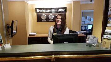 A reception desk at Buckatree Jail Hotel with a sign displaying clocks for New York and London, a computer, and a jar labeled "Monthly Business Card Draw".