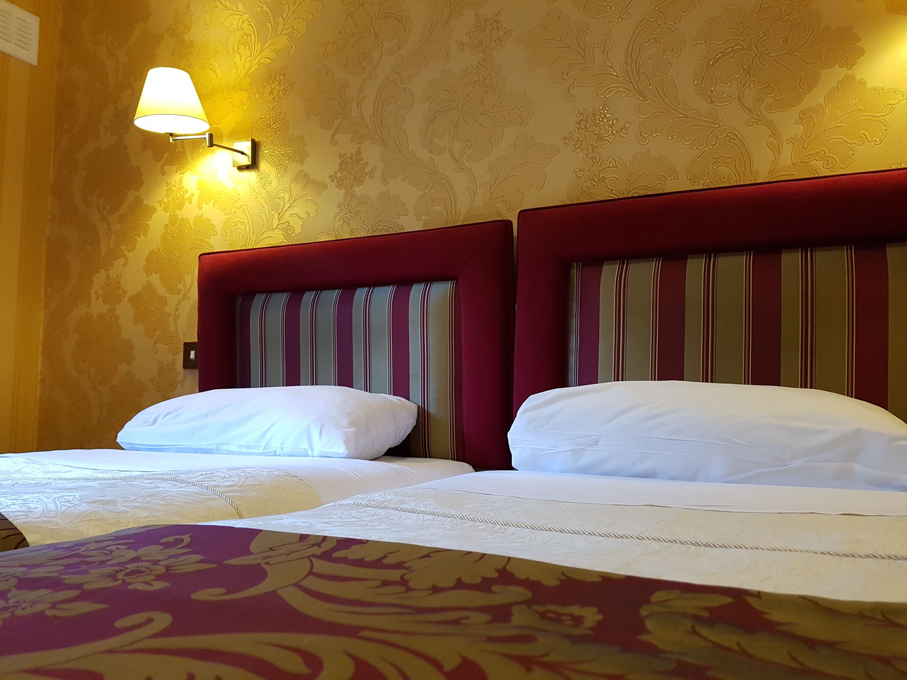 Habitación de hotel con dos camas, cada una con cabecero a rayas, una lámpara de pared y una alfombra estampada.