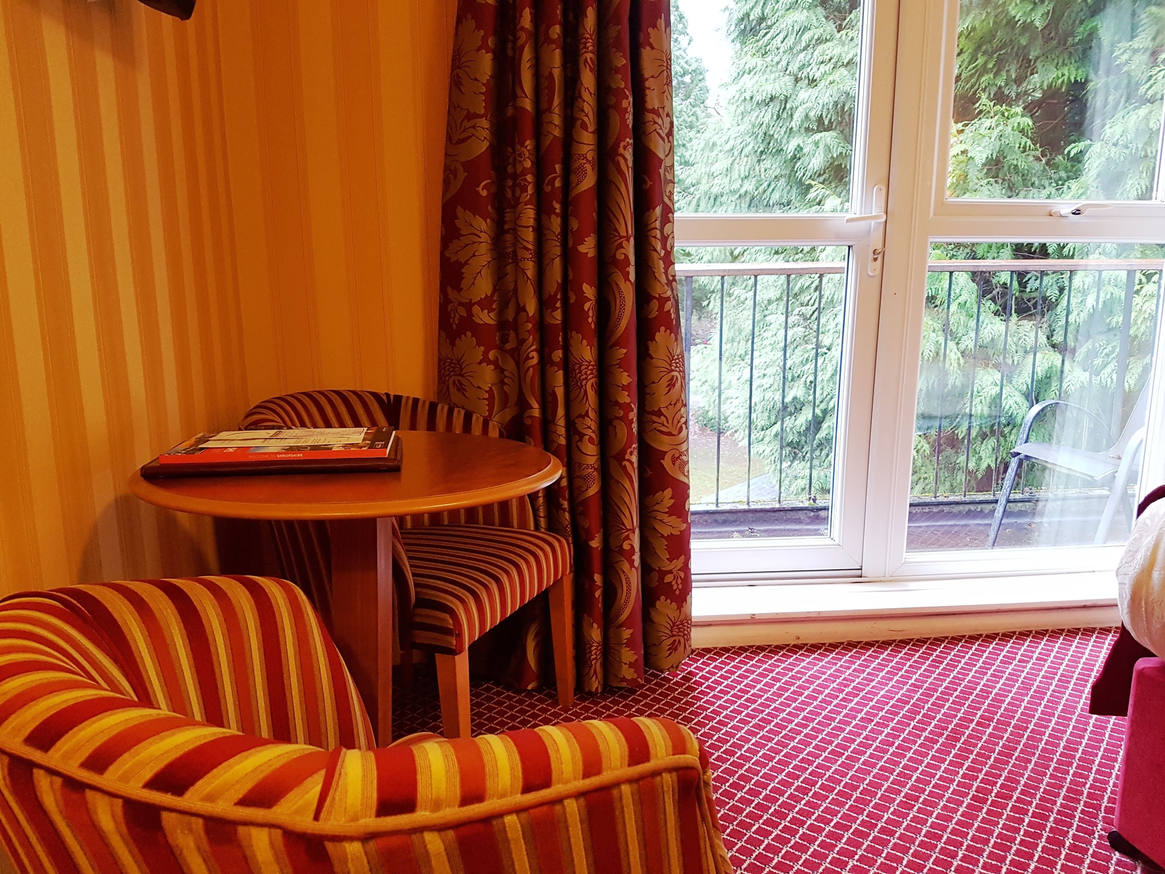 A room with a round table, two striped chairs, a red patterned sofa, and a large window with a view of greenery outside.