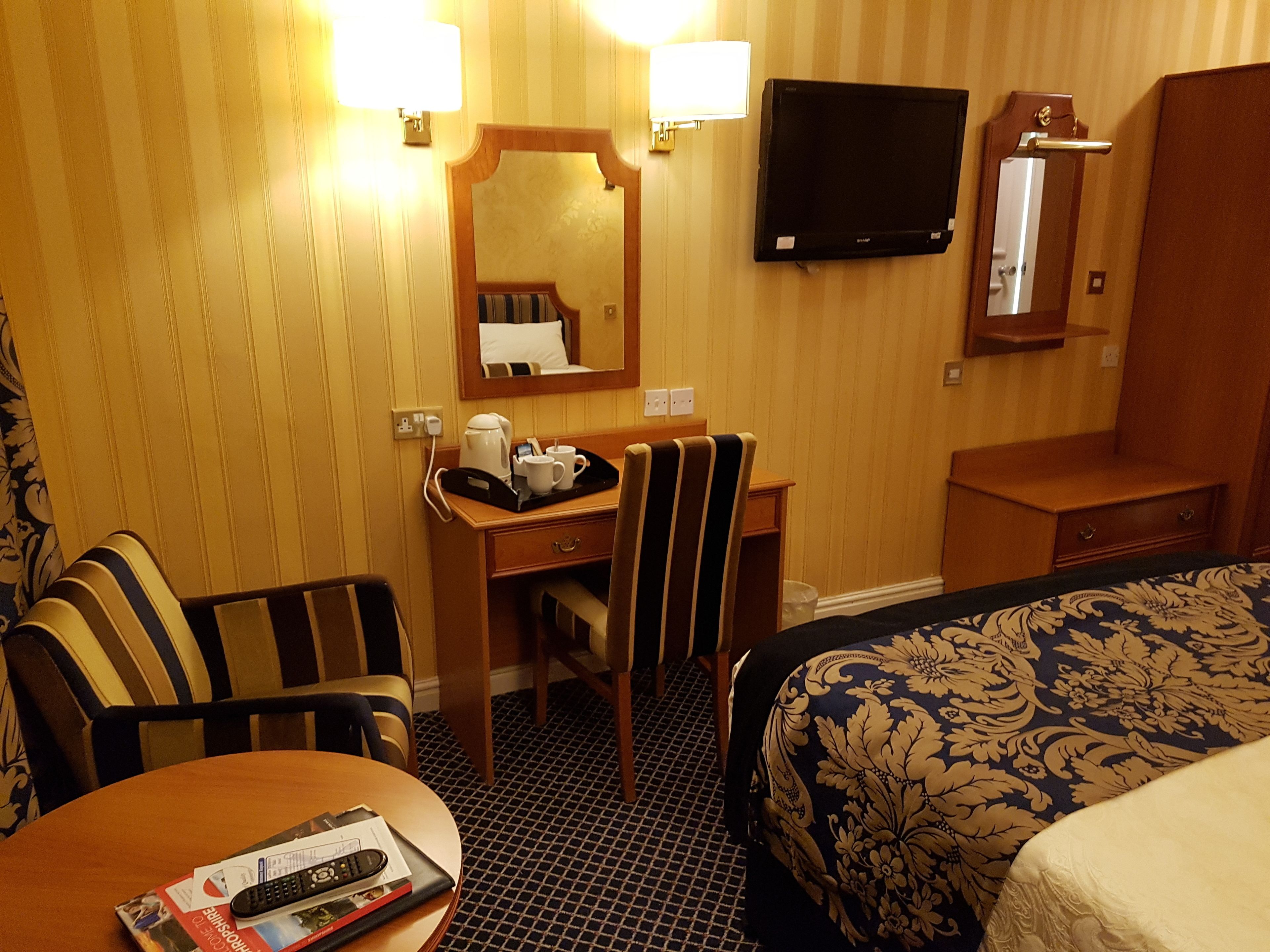 A hotel room with a bed, a desk with a chair, a television mounted on the wall, and a mirror above the desk.
