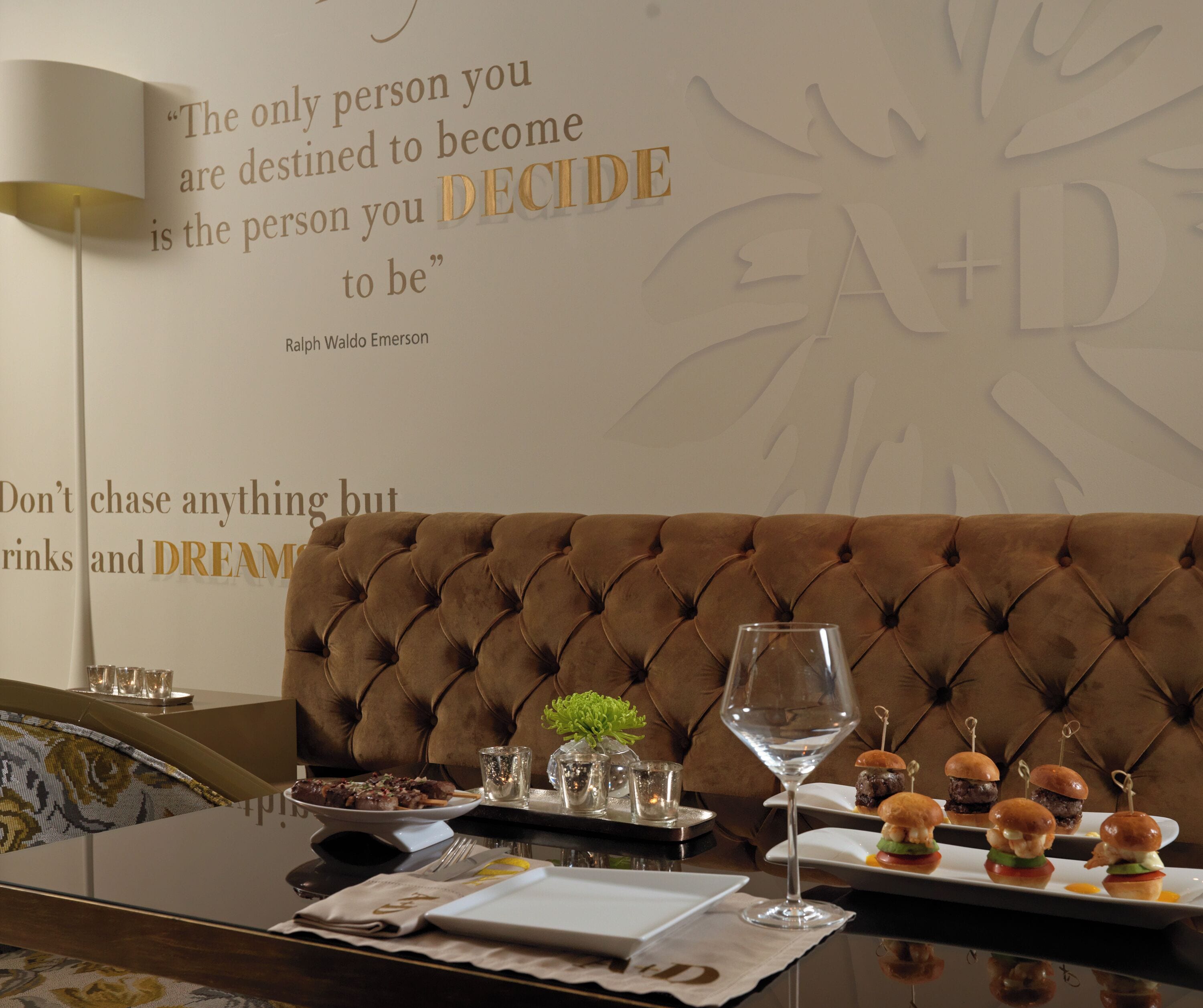 A dining area with a brown tufted sofa, a glass wine glass, and a tray of mini burgers.