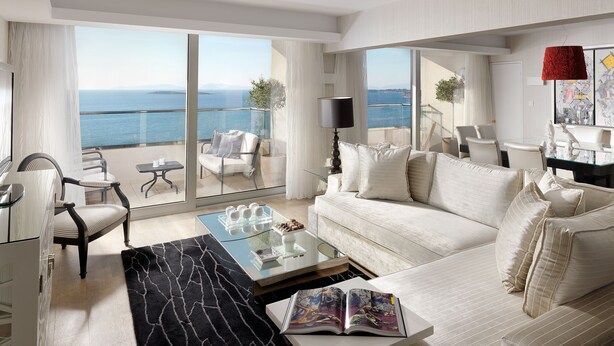 A spacious living room with a large sofa, a coffee table, and a view of the sea through floor-to-ceiling windows. - Divani Apollon Palace & Thalasso (Vari-Voula-Vouliagmeni)