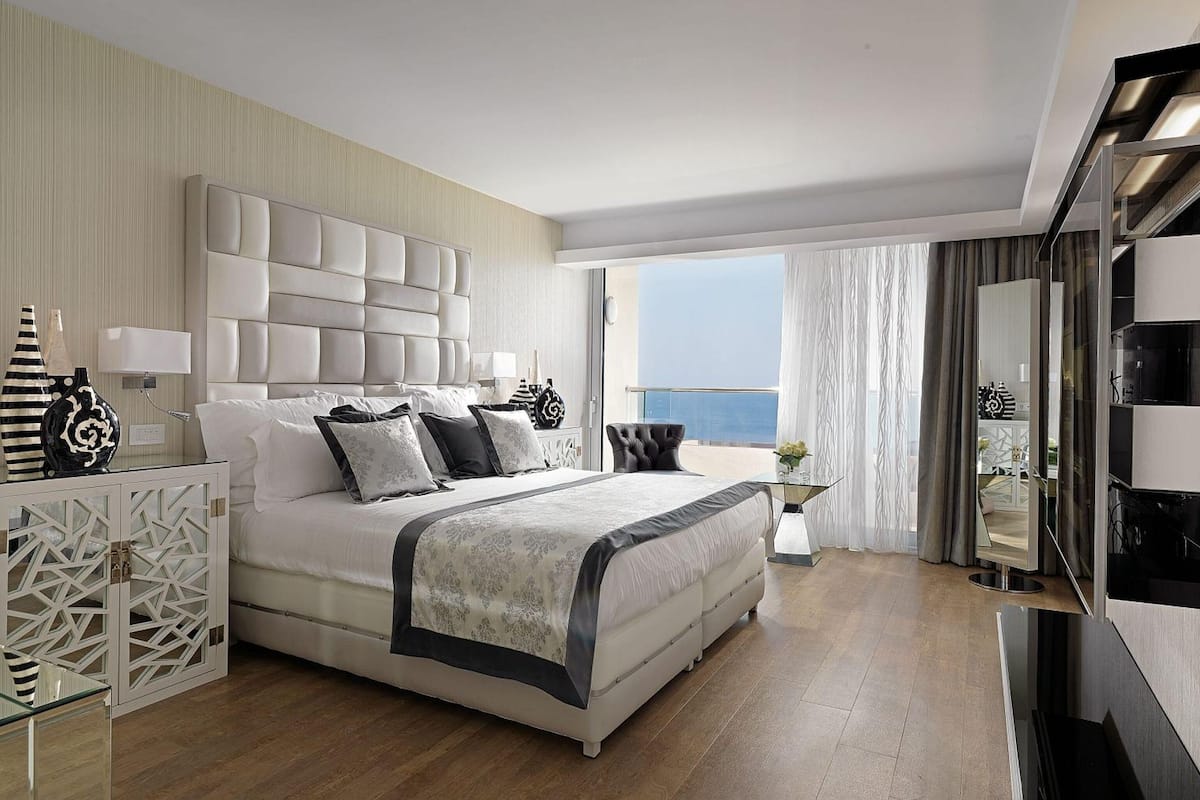 A modern hotel room with a large bed, a balcony with a view, a TV, and a minibar.