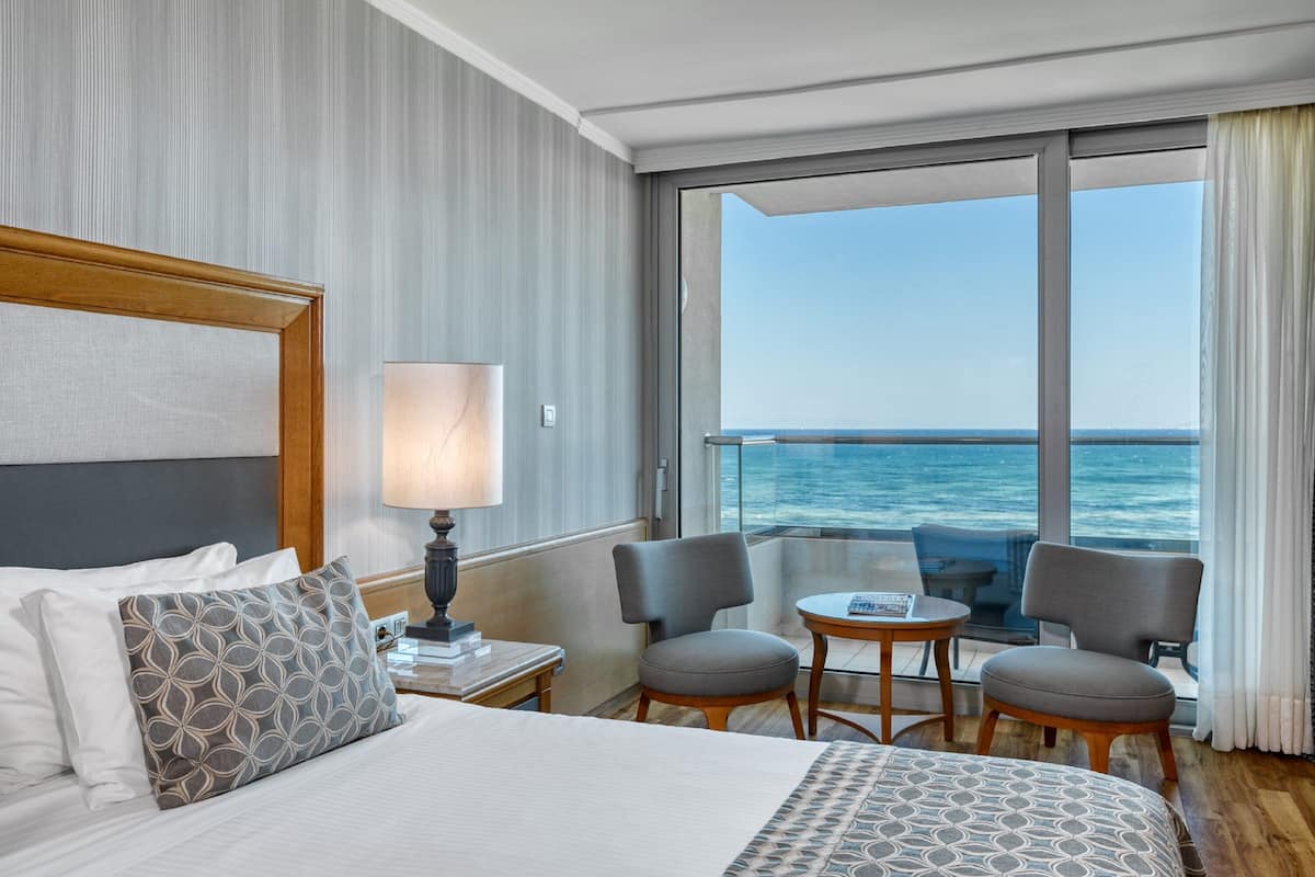 A hotel room with a large bed, a nightstand with a lamp, two armchairs, a small table, and a view of the ocean through large windows.