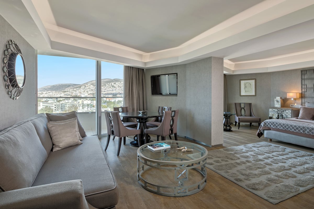 A modern hotel room with a sofa, a glass coffee table, a dining area with chairs, a TV mounted on the wall, and a bed with a patterned bedspread.
