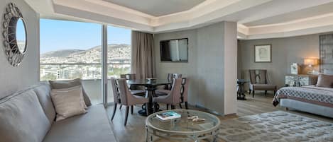Una habitación de hotel moderna con sofá, mesa de centro de vidrio, zona de comedor con sillas, televisor montado en la pared y cama con colcha estampada.