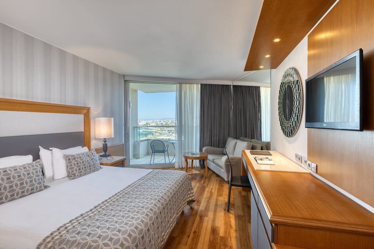 A hotel room with a large bed, a desk, a chair, a TV, and a balcony with a view.