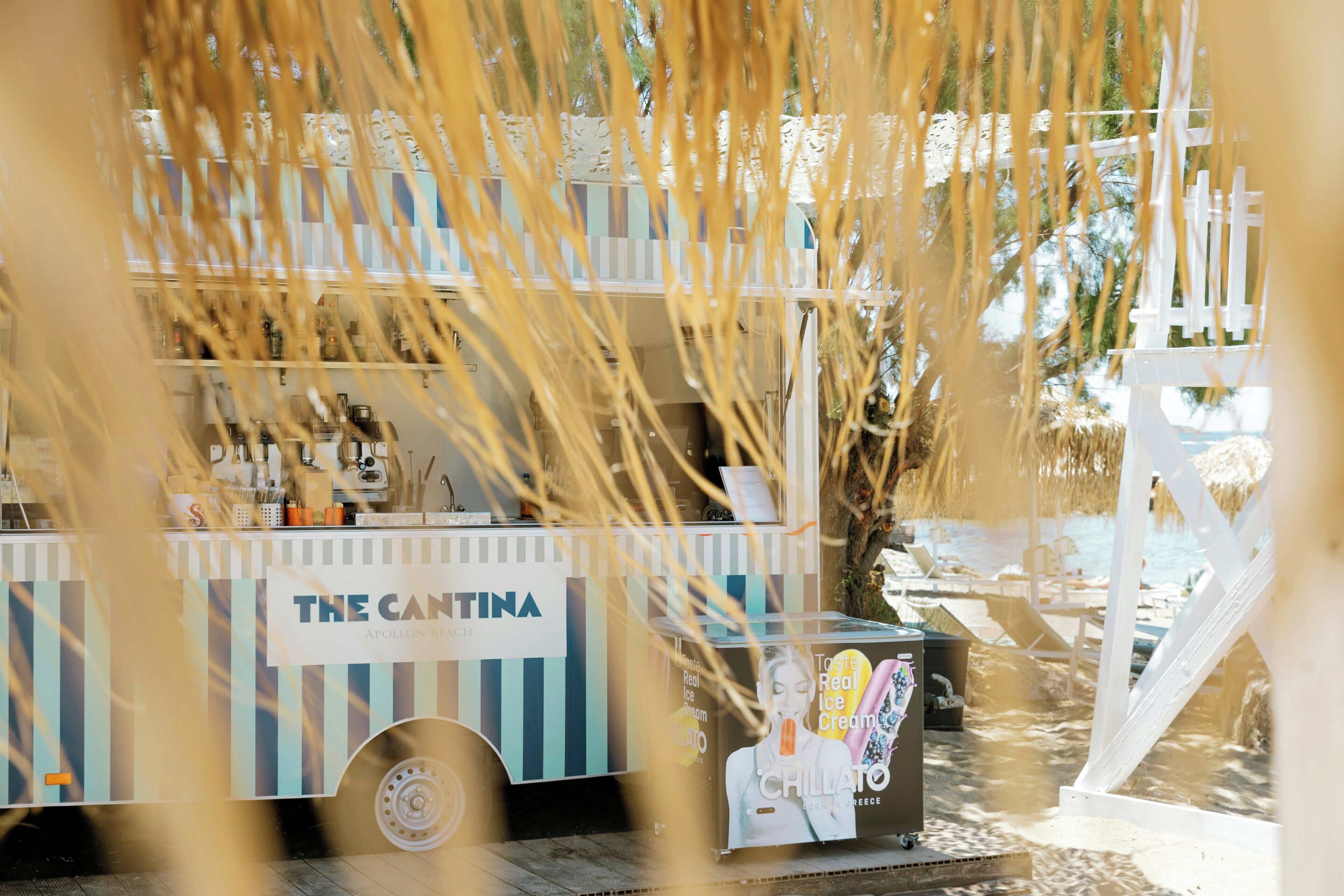 A food truck with a sign reading 'The Cantina' parked near a beach with lounge chairs and a palm tree.