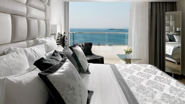 A modern hotel room with a large bed, a view of the sea, and a balcony with a glass table. - Divani Apollon Palace & Thalasso (Vari-Voula-Vouliagmeni)
