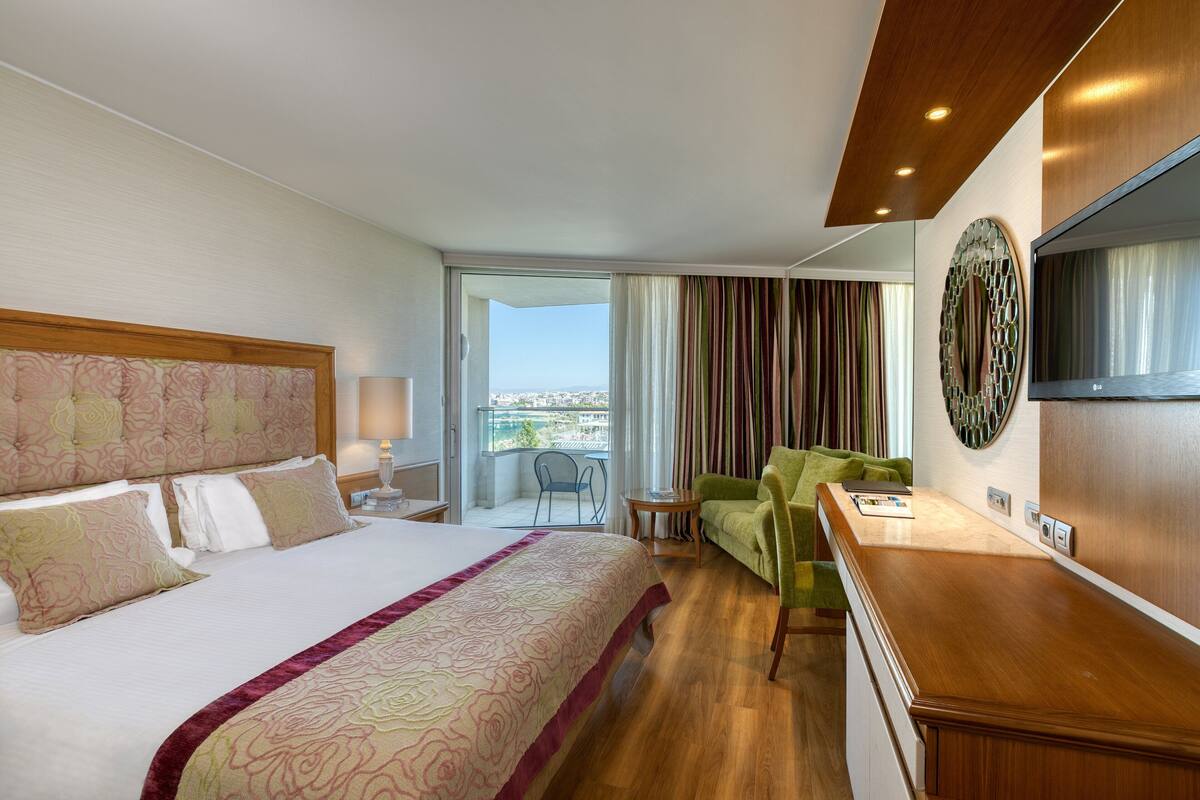 A hotel room with a large bed, a desk, a chair, a TV, and a balcony with curtains.