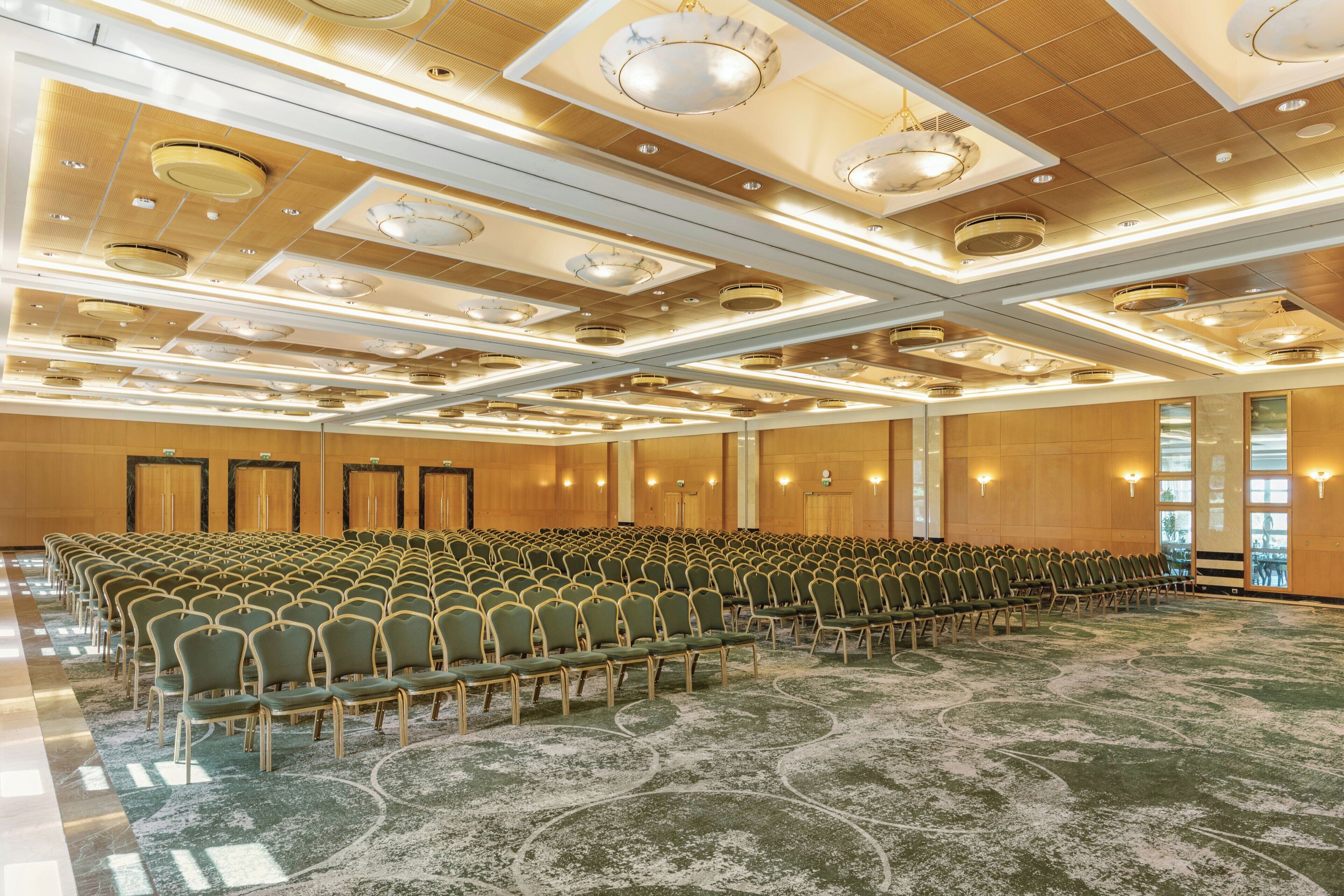 Une salle de conférence avec des rangées de chaises, une moquette à motifs et un plafond doté d’un éclairage encastré et d’éléments décoratifs.