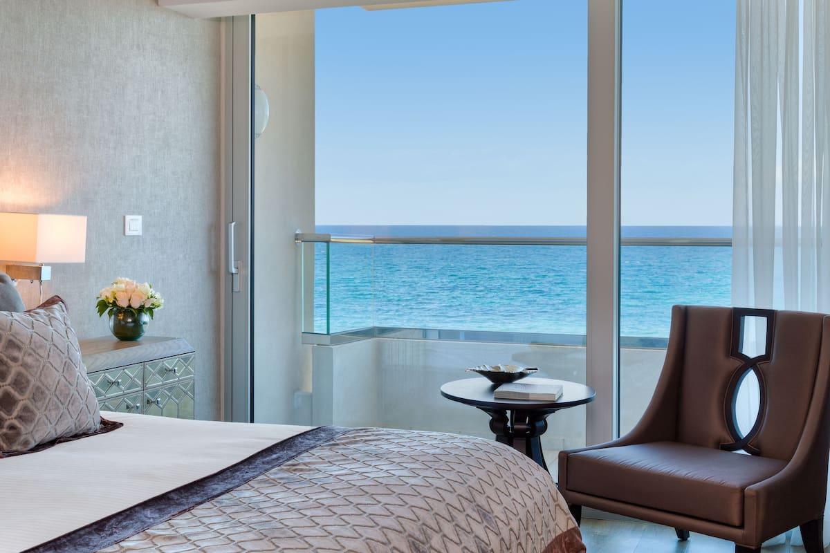 A hotel room with a bed, a chair, a small table, and a view of the ocean.