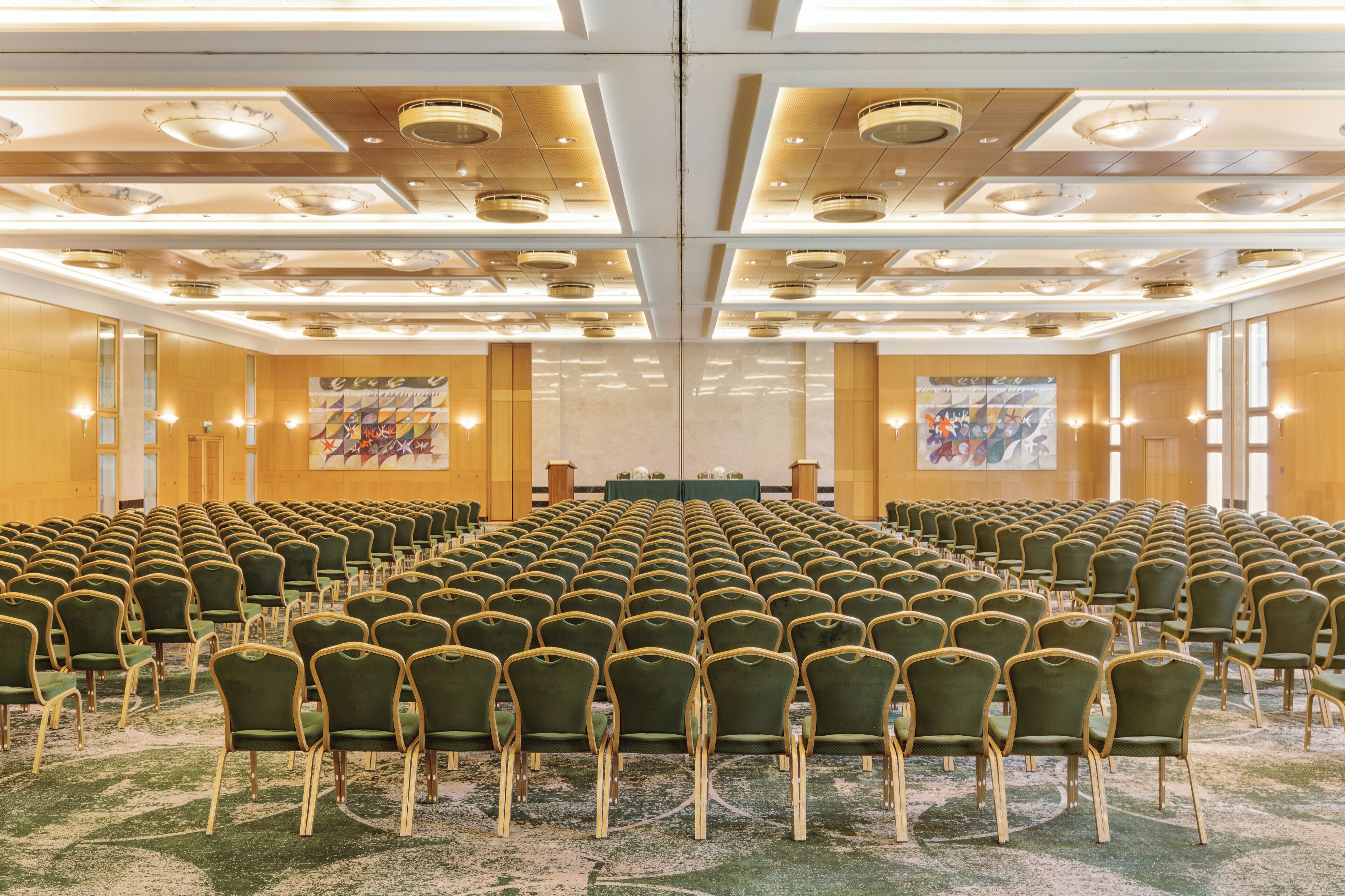 A conference room with rows of green chairs, a stage with a table, and a large poster on the wall.
