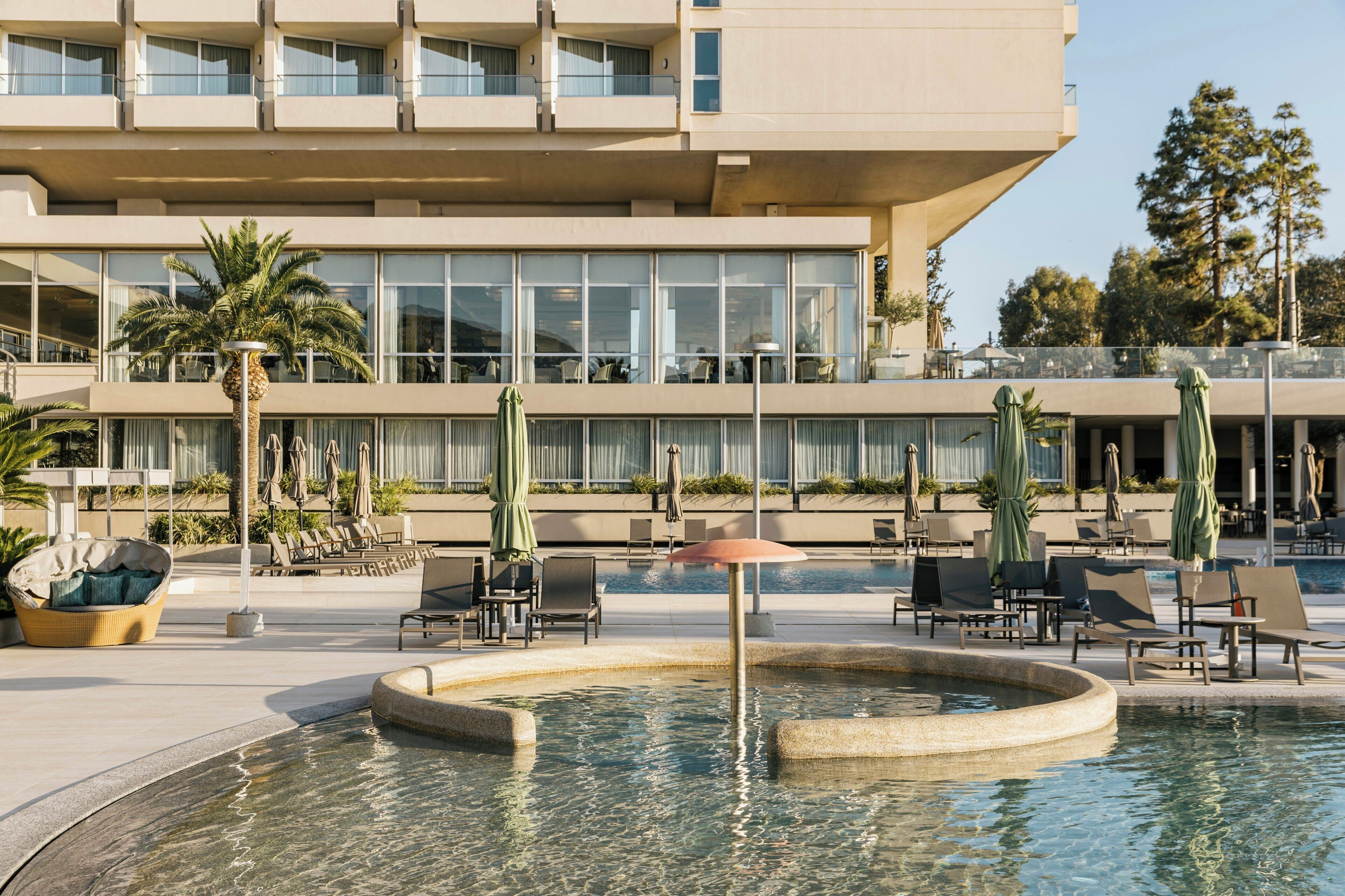 Un hotel moderno con zona de piscina, mobiliario exterior y un edificio grande de varios pisos.