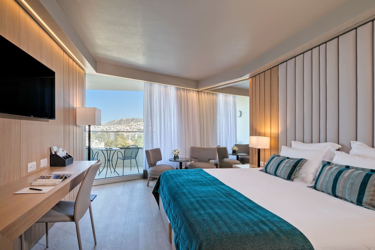 A hotel room with a large bed, a desk with a chair, a TV, and a balcony with a view.