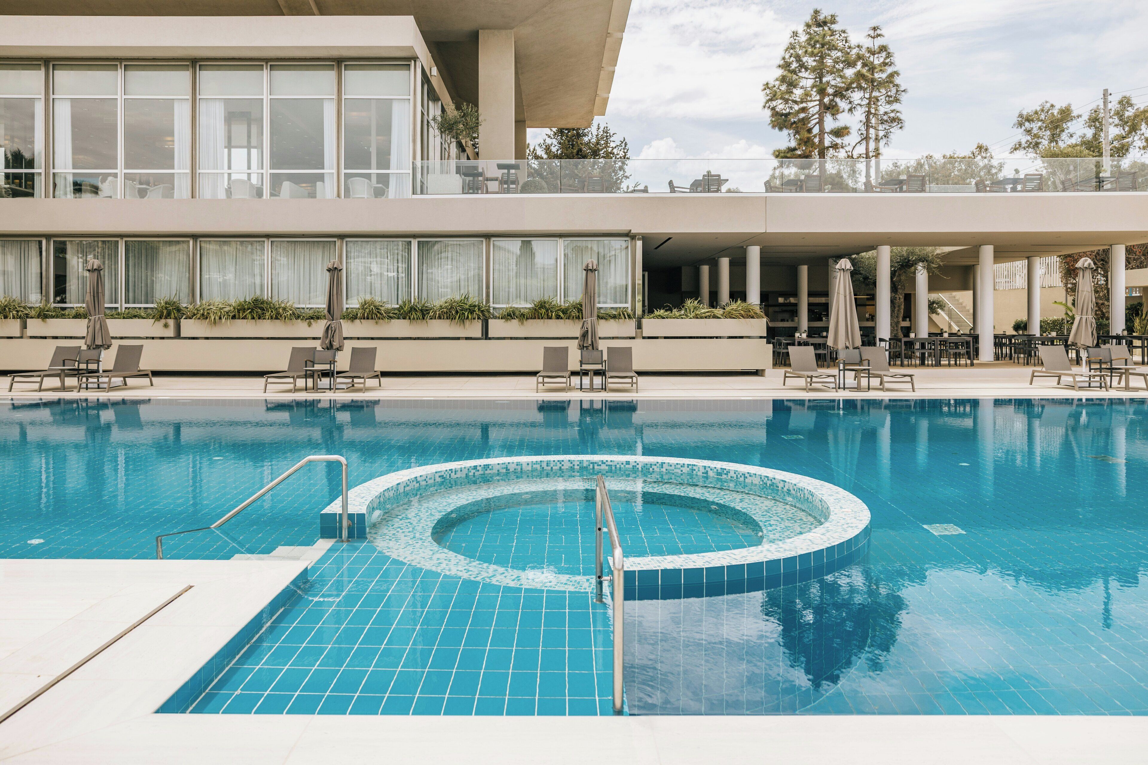 A modern hotel pool area with a central hot tub, lounge chairs, and a covered seating area.