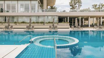 A modern hotel pool area with a central hot tub, lounge chairs, and a covered seating area.