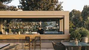 An outdoor bar area with a bartender, wooden chairs, and a glass table. - Divani Apollon Palace & Thalasso (Vari-Voula-Vouliagmeni)