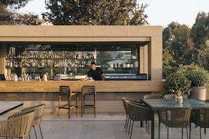 An outdoor bar area with a bartender, wooden chairs, and a glass table. - Divani Apollon Palace & Thalasso (Vari-Voula-Vouliagmeni)