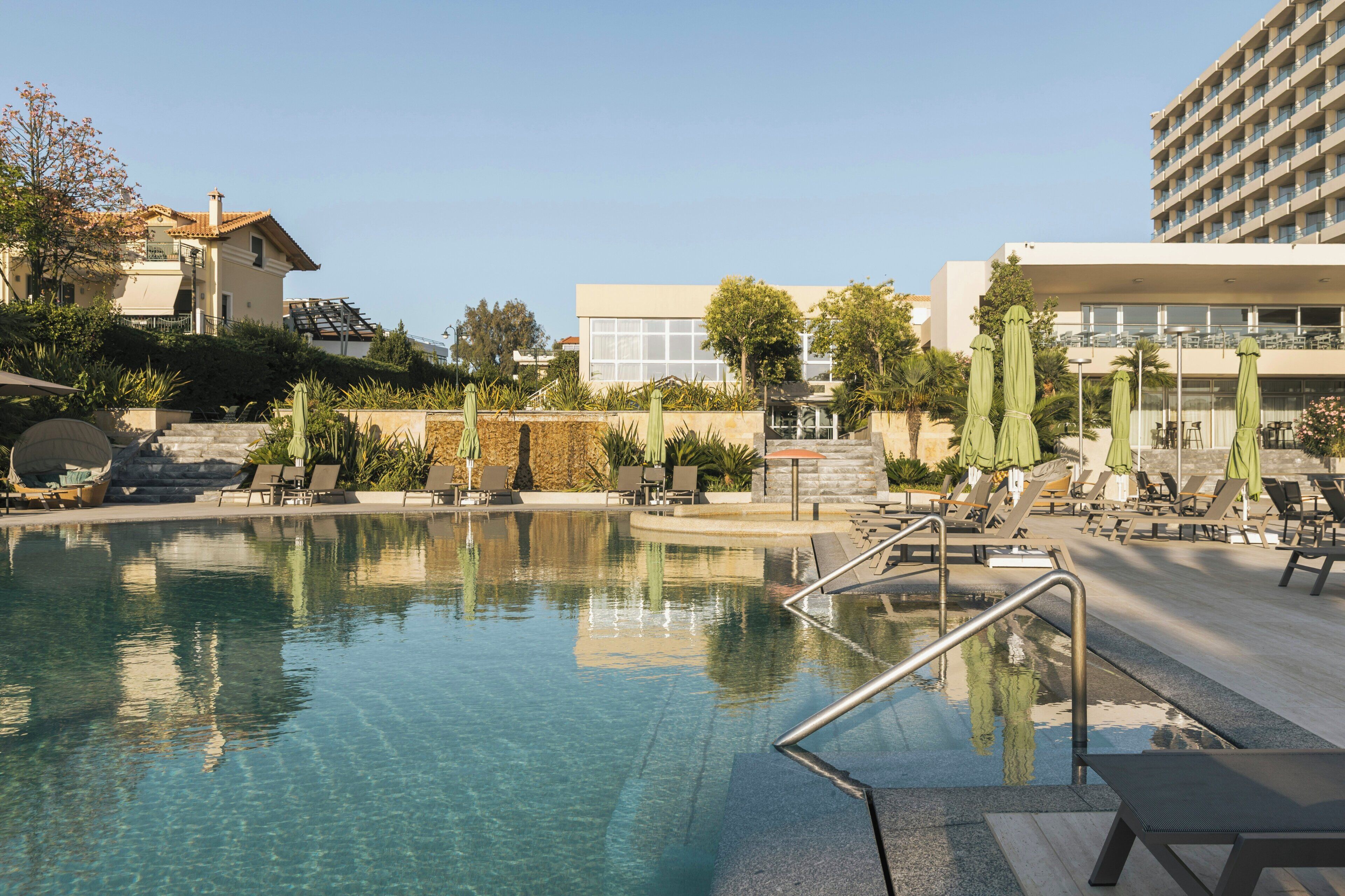 A swimming pool with a clear blue water surface, surrounded by lounge chairs and umbrellas, and adjacent to a building with a modern architectural style.