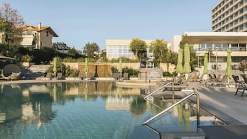 A swimming pool with a clear blue water surface, surrounded by lounge chairs and umbrellas, and adjacent to a building with a modern architectural style.