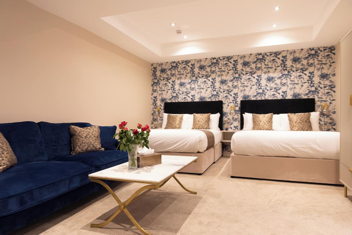 Ein Hotelzimmer mit einem blauen Sofa, zwei Betten mit schwarzen Kopfteilen, einem weißen Couchtisch und floralem Wandpapier.