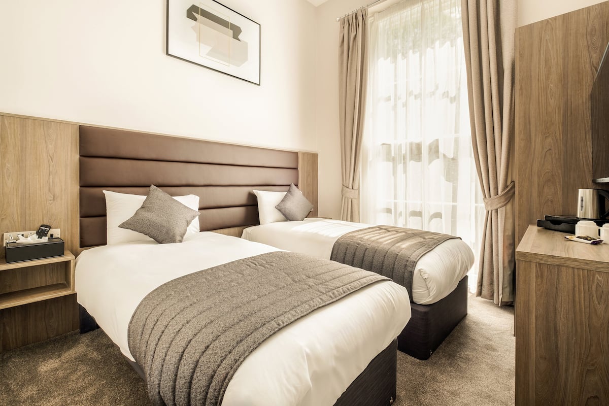 Ein Hotelzimmer mit zwei Betten, einem Holzkopfteil, einem Schreibtisch und einem Fenster mit Vorhängen.