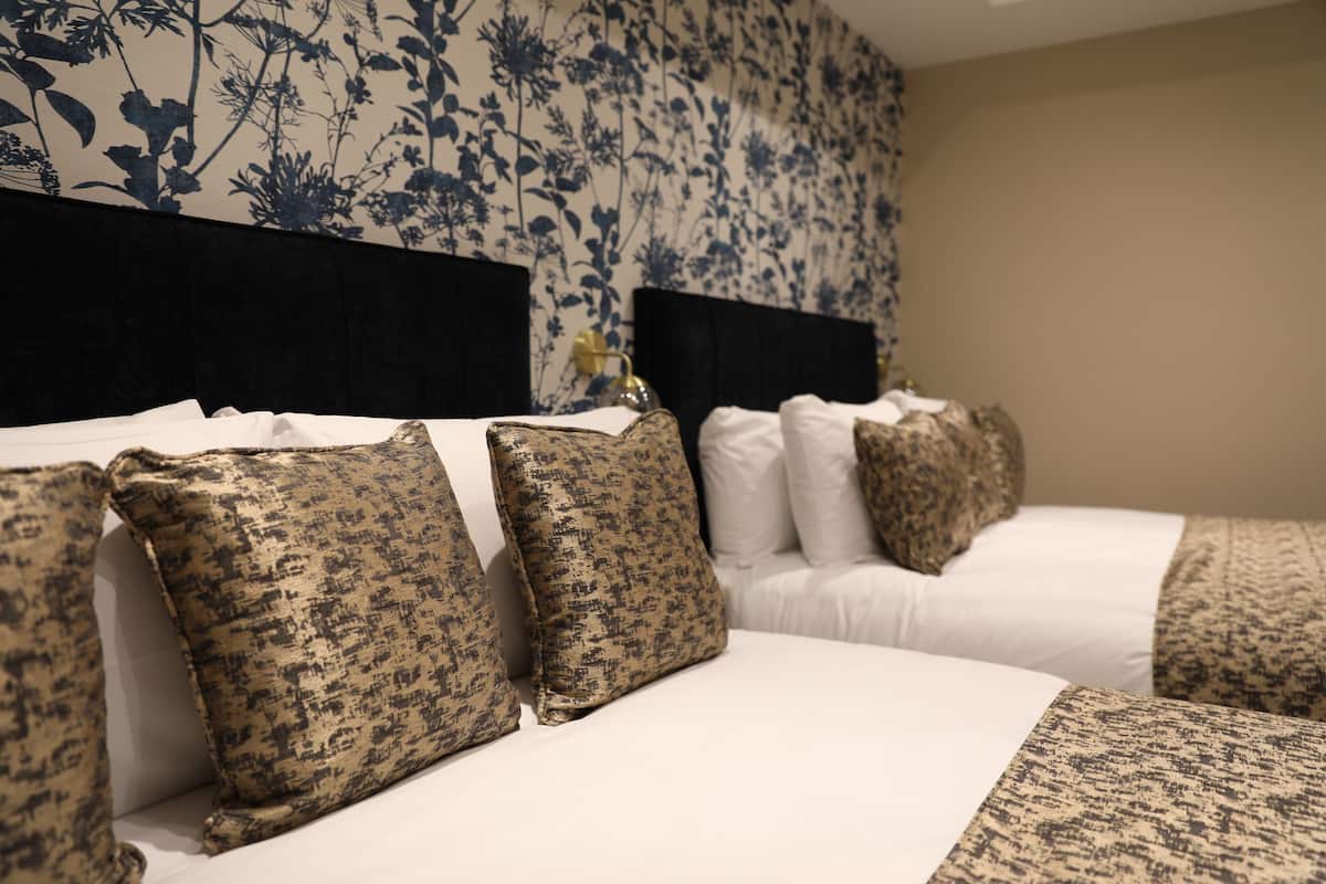 Ein Hotelzimmer mit floralem Wandpapier, einem schwarzen Kopfteil und mehreren Betten mit gemusterten Kissen.