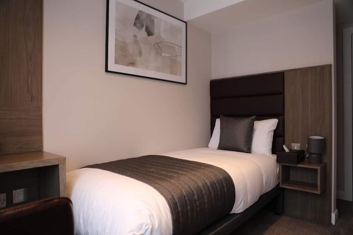 Ein modernes Hotelzimmer mit einem Einzelbett, einem Holzkopfstück, einem Nachttisch mit Lampe und einem gerahmten abstrakten Kunstwerk an der Wand.