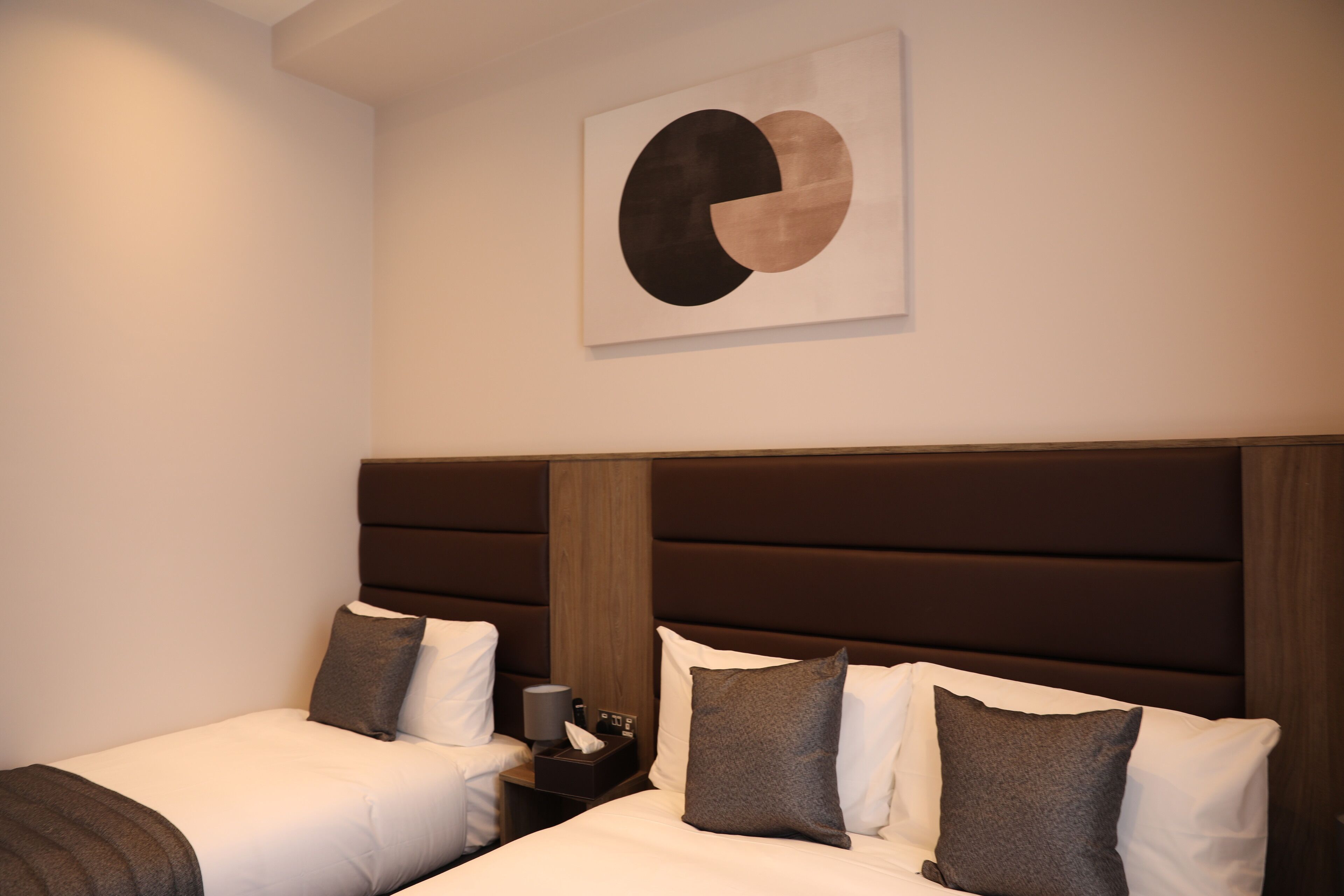 Hotellihuone, jossa on kaksi sänkyä, suuri abstrakti maalaus seinällä ja puinen pääty.