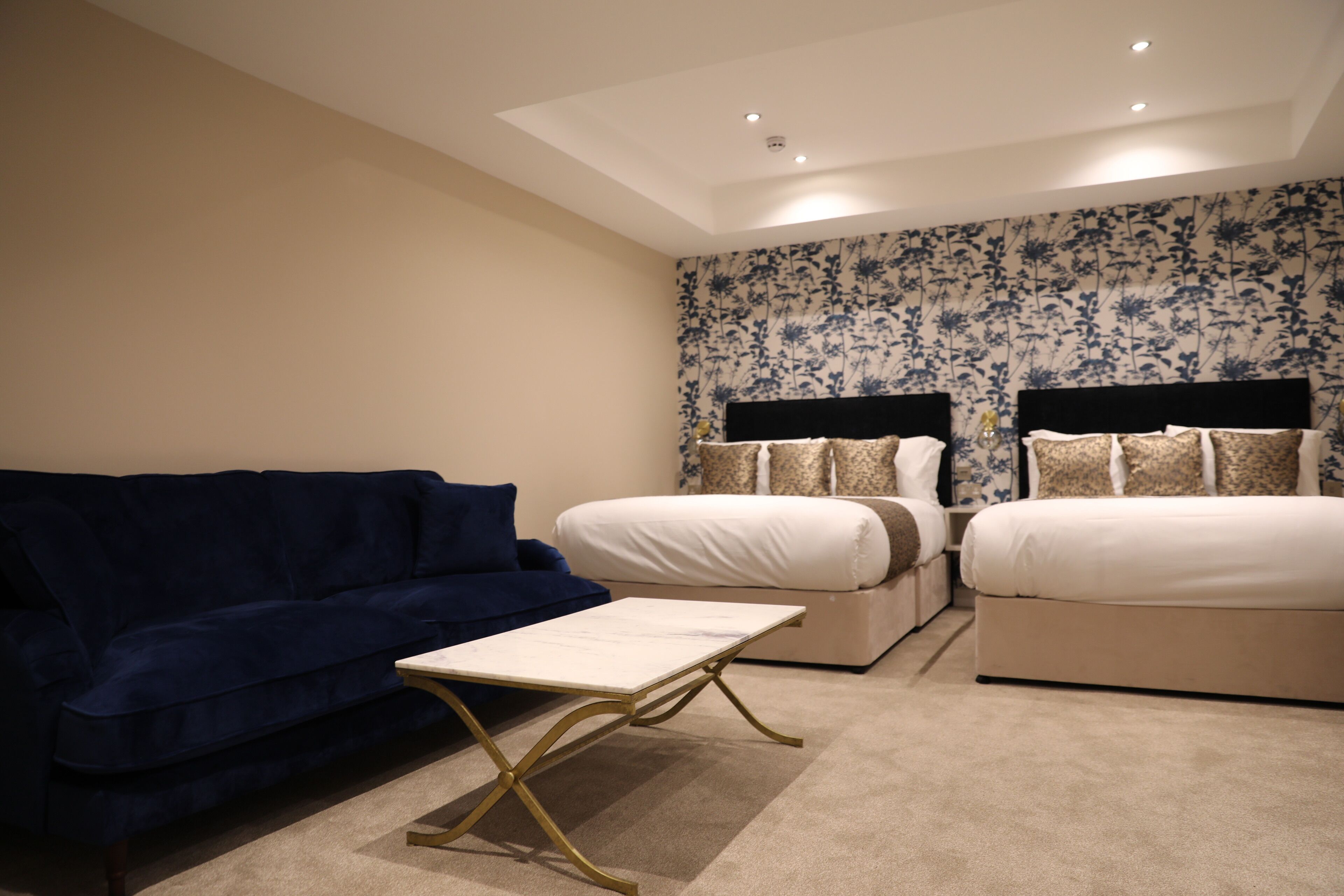 A hotel room with a blue sofa, a small table, two beds with white bedding, and a floral wallpaper.