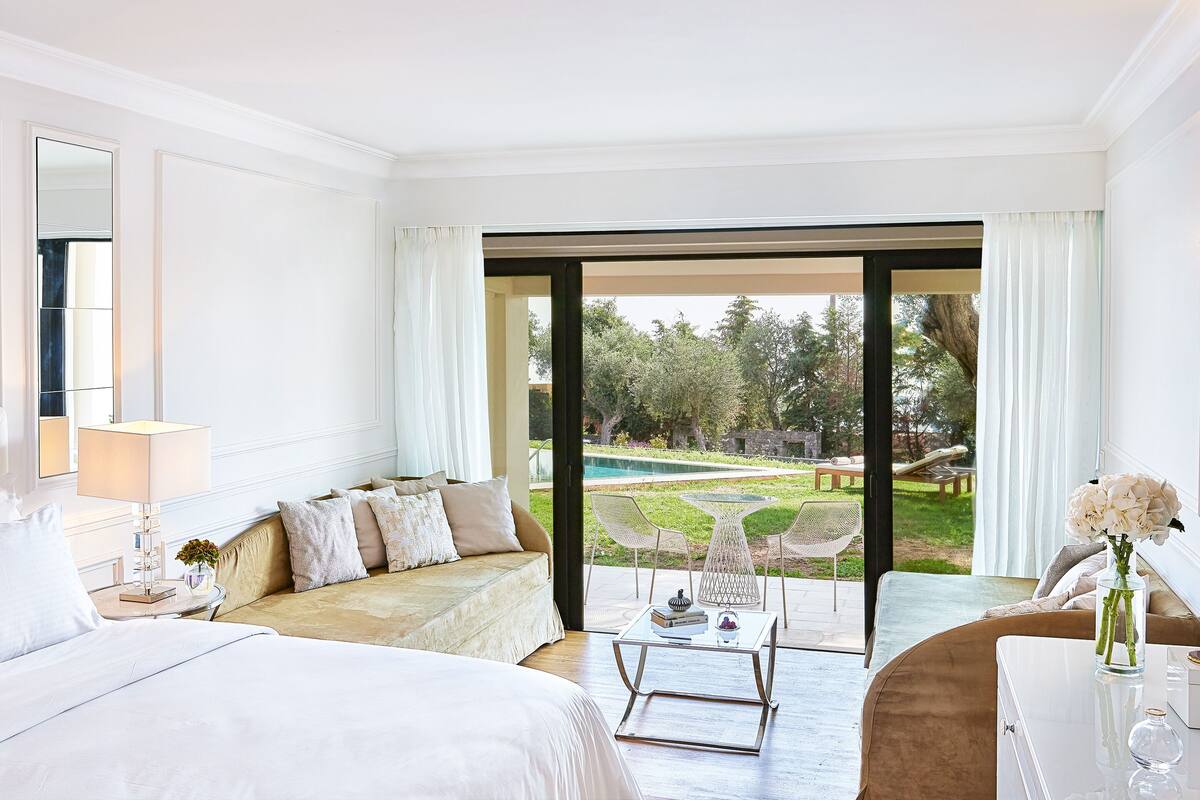 A hotel room with a large bed, a sofa, a dining table, and a view of a garden and pool.