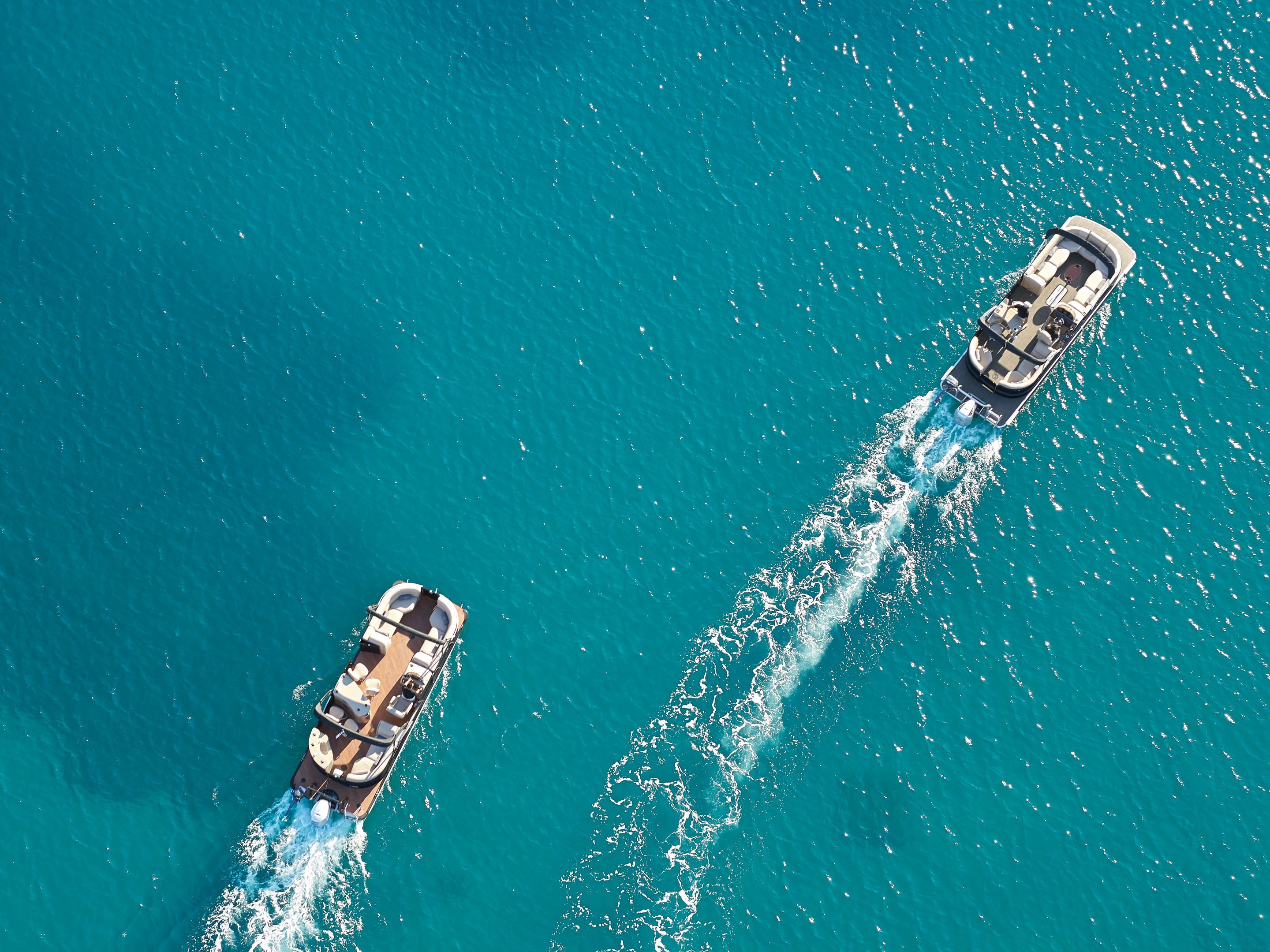 Two boats on a turquoise sea, one smaller and one larger, both with seating and wake trails.