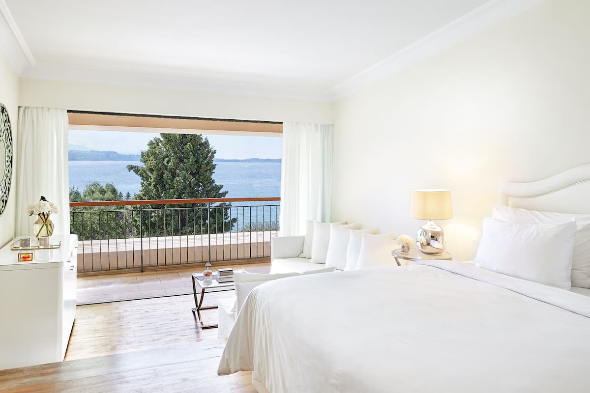 A bedroom with a large bed, a nightstand, a lamp, and a view of the sea through a balcony.