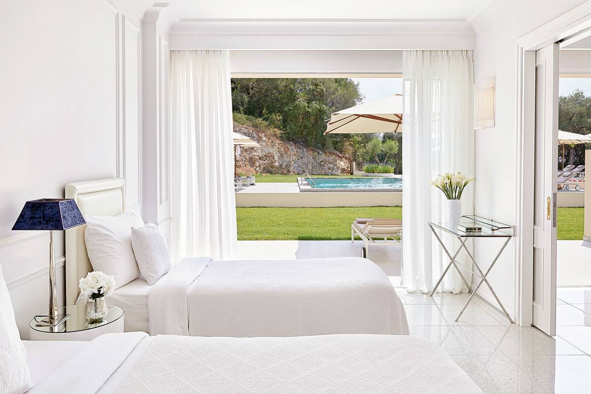 A spacious bedroom with a large bed, a bedside table with a lamp, and a view of a swimming pool and outdoor seating area.