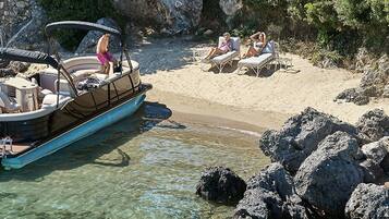 A boat docked near a rocky shoreline with clear water. There are lounge chairs and a small table on the beach.