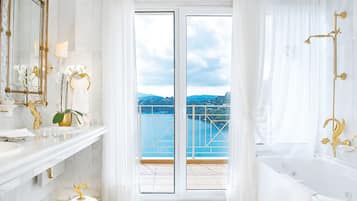 Une salle de bain luxueuse dotée d’une grande baignoire, d’un meuble-lavabo avec des accessoires dorés et offrant une vue sur un balcon surplombant une étendue d’eau.