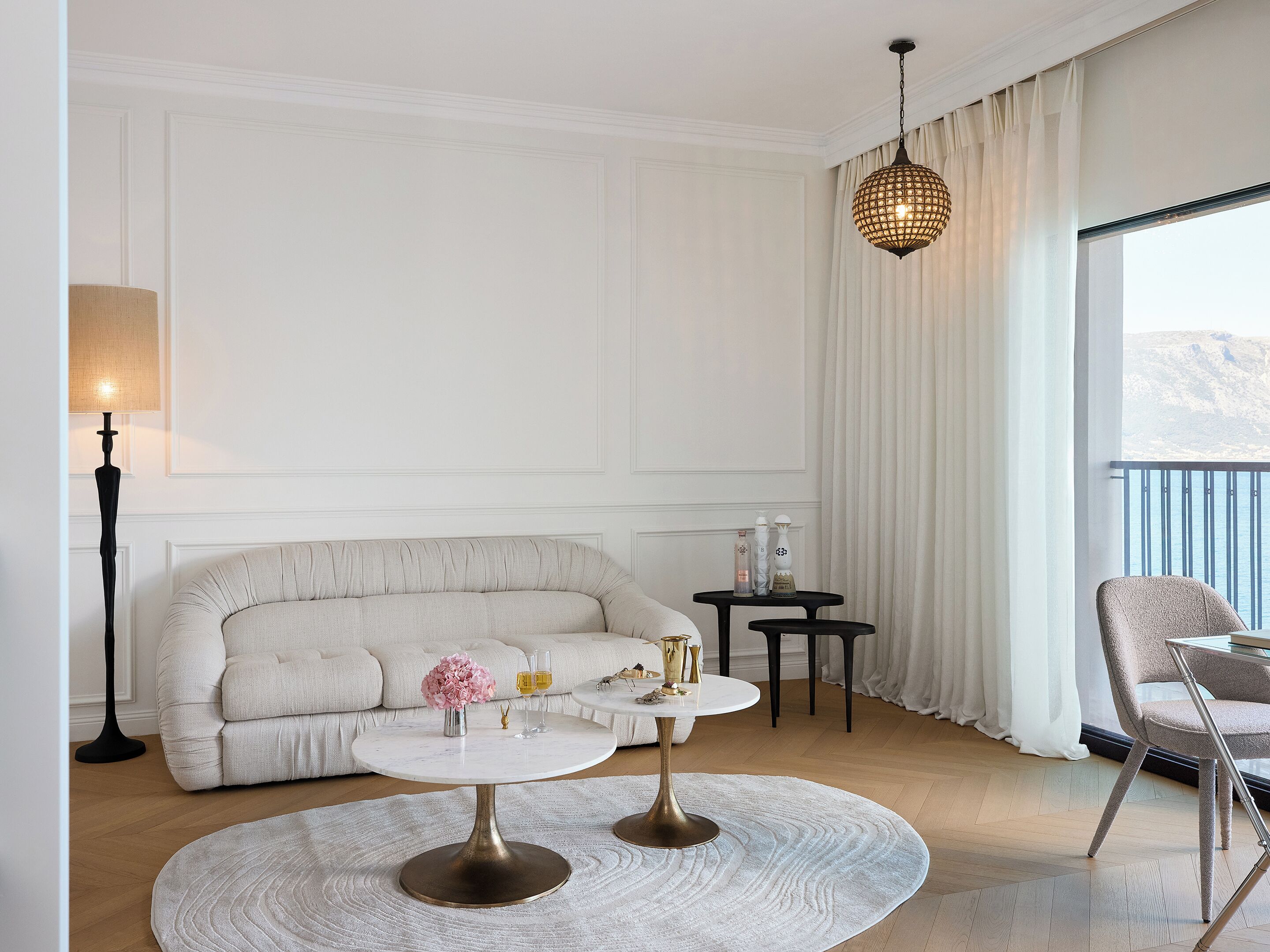 Uma sala de estar moderna com um sofá bege, uma mesa de centro redonda, um abajur e vista para o mar através de grandes janelas.