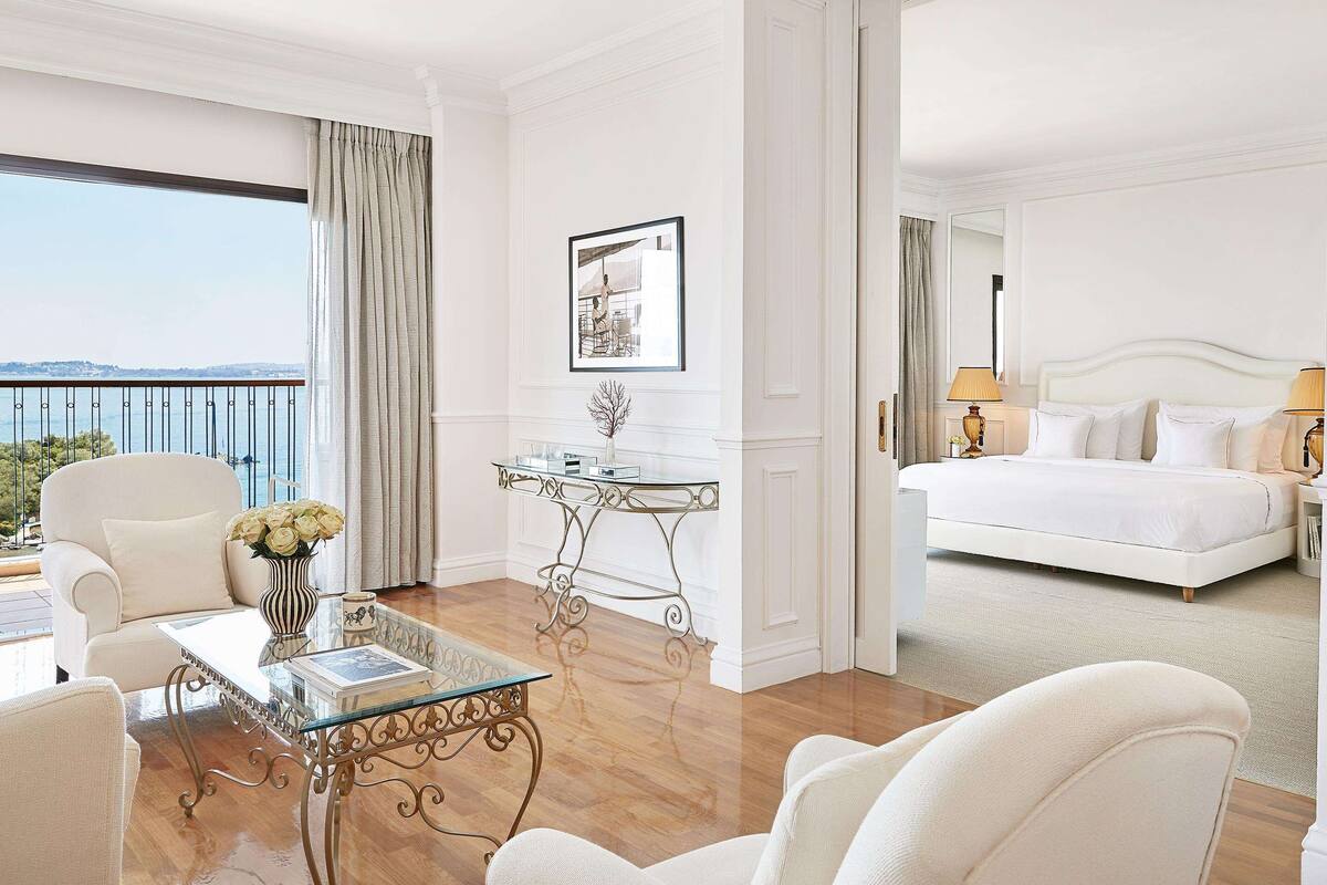 A hotel room with a balcony, a bed, a sitting area with a glass table, and a white color scheme.