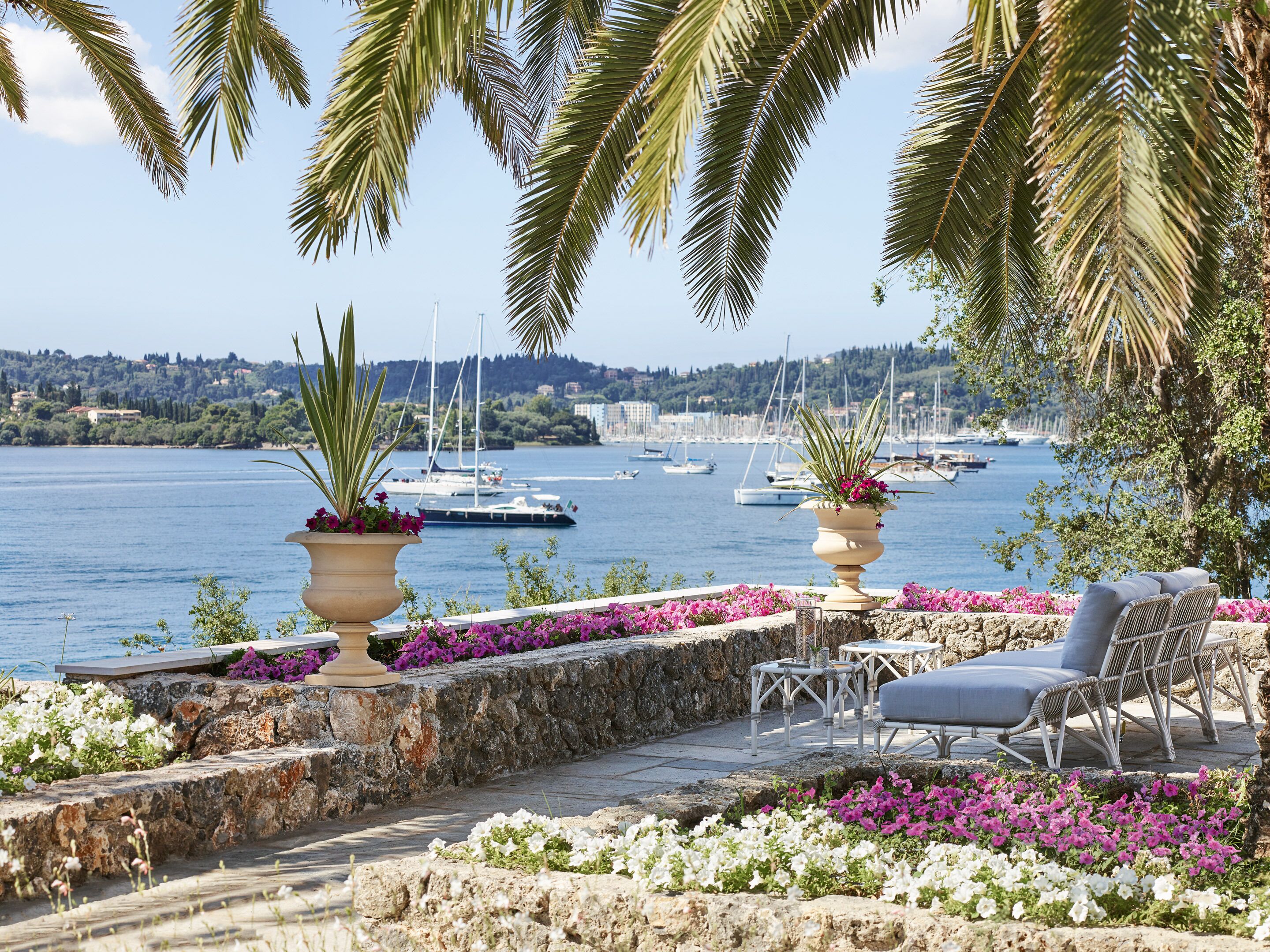 Una terraza costera con muros de piedra, plantas en macetas y mobiliario exterior, con vistas a un puerto deportivo con barcos.