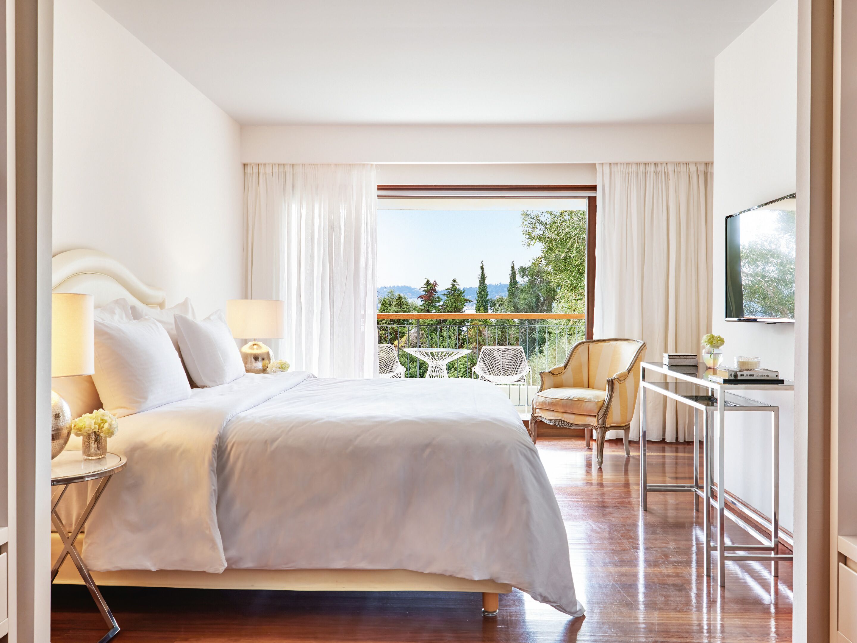 A hotel room with a large bed, a desk with a chair, a TV, and a balcony with outdoor seating.