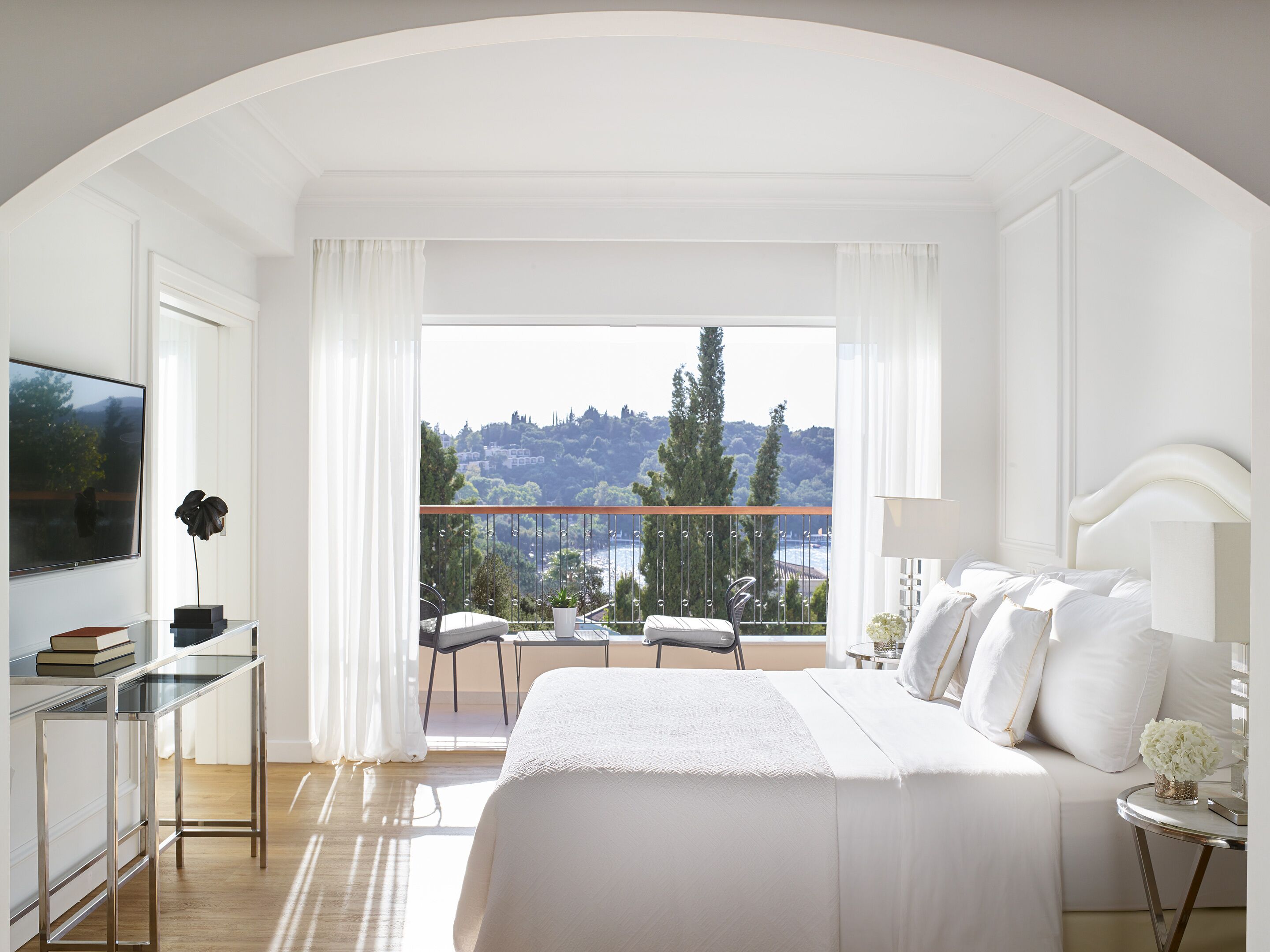 Een ruime slaapkamer met een groot bed, een televisie, een balkon met uitzicht op de stad en een zithoek met een tafel en stoelen.