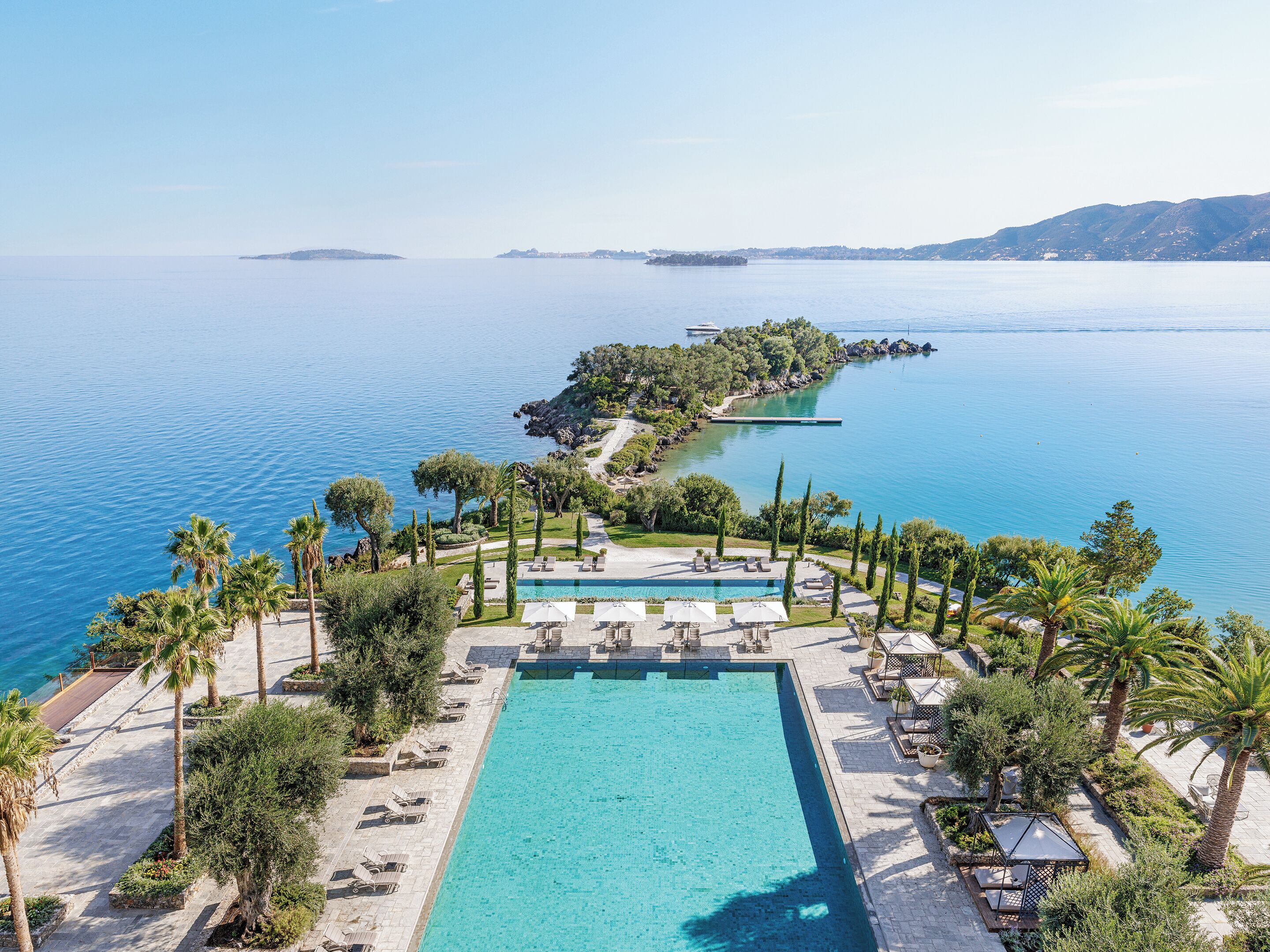 A resort with a large pool, sun loungers, palm trees, and a view of the sea.