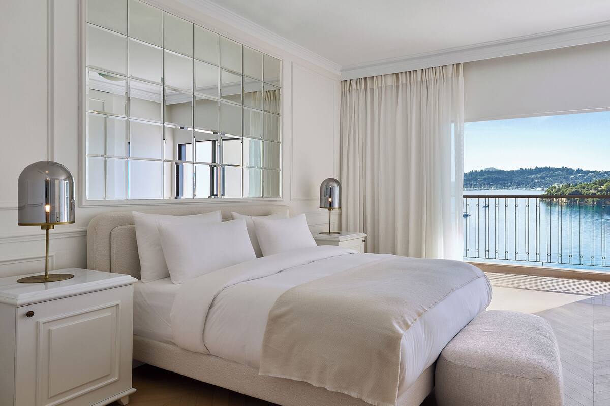 A bedroom with a large bed, a nightstand with a lamp, a mirrored wardrobe, and a balcony with a view of the water.