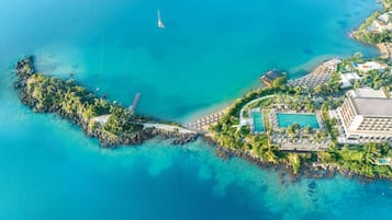 Vista aérea de una isla resort con piscina central, un puente que la conecta a tierra firme y diversos edificios y vegetación.