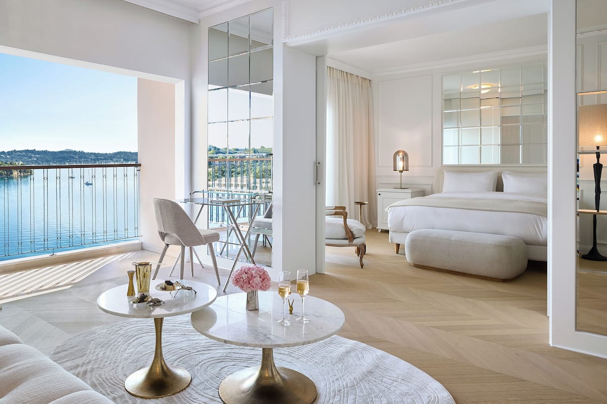 A modern hotel room with a balcony, a bed, a sitting area with a table and chairs, and a view of the water.