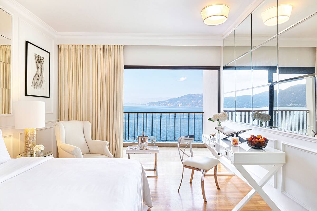 A hotel room with a bed, a desk with a chair, a large window with a view of the sea, and a balcony with a railing.